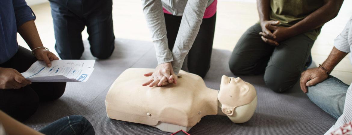 CPR dummy in center of room with hands applied, people in circle around it