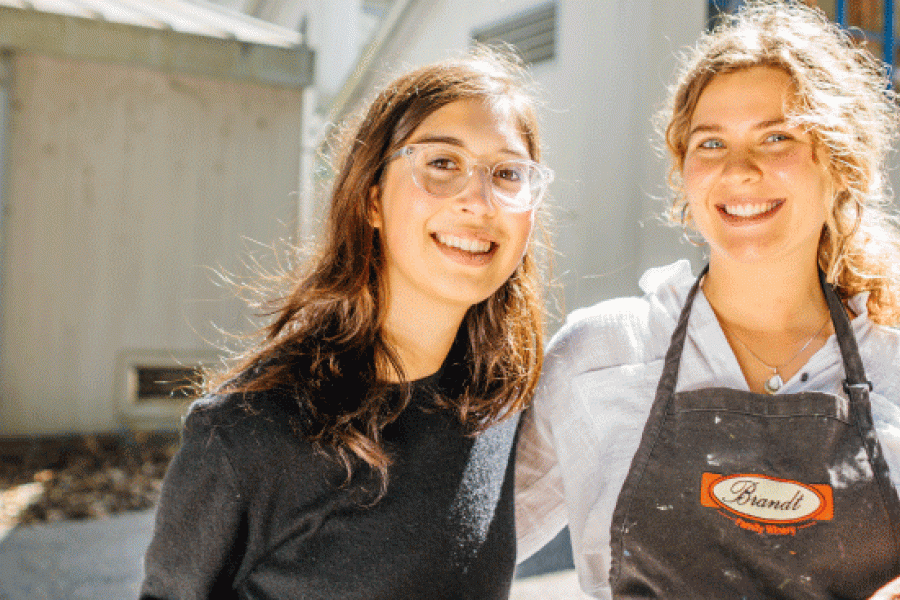 Photo of two female students outside a building wearing painting clothes and carrying a canvas